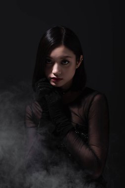 portrait of young asian woman in gothic outfit and gloves on grey with smoke
