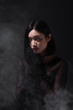 portrait of young asian woman in gothic outfit and gloves looking away on grey with smoke