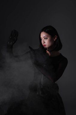 brunette asian woman in gothic outfit and gloves gesturing on grey with smoke