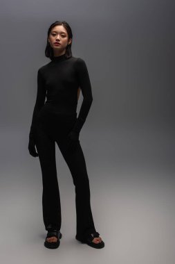 full length of stylish asian woman in total black outfit standing on grey 