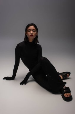 full length of stylish asian woman in total black outfit sitting on grey 