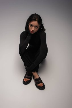top view of pretty and young asian woman in total black outfit sitting on grey 