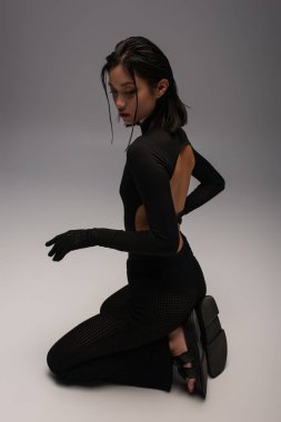 full length of young asian woman in total black and stylish outfit sitting on knees on grey background 