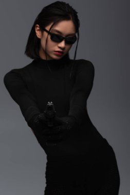dangerous asian woman in total black outfit and stylish sunglasses aiming with gun isolated on grey