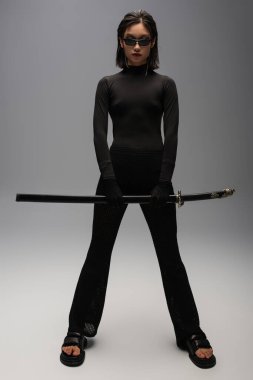 full length of asian woman in black outfit and stylish sunglasses holding katana sword on grey