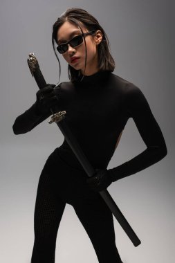 asian woman in black outfit and stylish sunglasses holding katana sword on grey