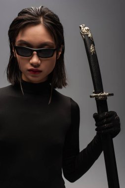 brunette asian woman in black clothes and stylish sunglasses holding katana sword in scabbard isolated on grey