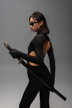 brunette asian woman in black outfit and stylish sunglasses posing with katana sword isolated on grey