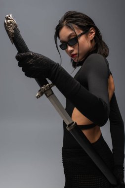 brunette asian woman in black outfit and stylish sunglasses drawing katana from scabbard isolated on grey