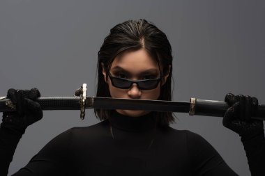 front view of asian woman in black outfit and stylish sunglasses pulling out katana from scabbard isolated on grey