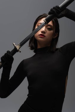 brunette asian woman in black outfit pulling out katana from scabbard isolated on grey