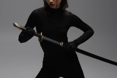 cropped view of woman in black outfit pulling out katana from scabbard isolated on grey clipart