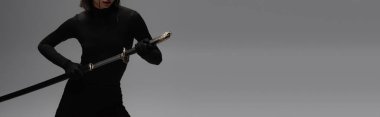 cropped view of woman in black outfit pulling out katana from scabbard isolated on grey, banner