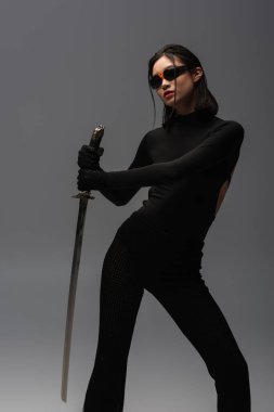 brunette asian woman in sunglasses and black clothes holding katana isolated on grey