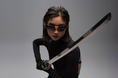 dangerous asian woman in sunglasses and black outfit holding katana isolated on grey