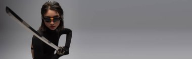brunette asian woman in sunglasses and black outfit holding katana isolated on grey, banner