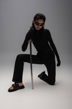 full length of dangerous asian woman in sunglasses and black clothes holding katana while standing on knee on grey