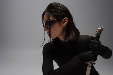 side view of dangerous asian woman in sunglasses and black outfit holding katana isolated on grey
