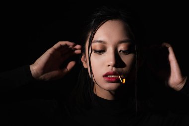 portrait of pretty asian woman with burning match in mouth posing isolated on black clipart