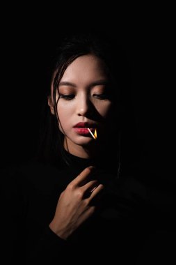 portrait of pretty asian model with burning matchstick in mouth isolated on black clipart
