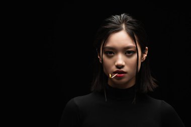 portrait of pretty asian woman with burning match in mouth looking at camera isolated on black