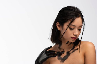 young asian woman with bare shoulders and hand in black paint touching body isolated on white