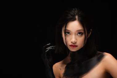 young asian woman with bare shoulders and hand in paint looking at camera isolated on black