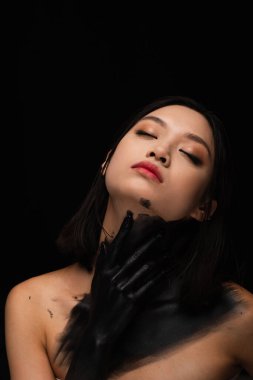 young asian woman with bare shoulders and hand in paint touching neck isolated on black