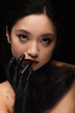 young asian woman with bare shoulders and hand in paint looking at camera while touching lip isolated on black
