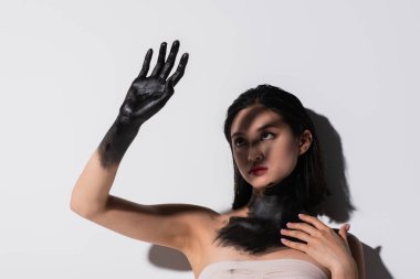 young asian woman holding hand in black paint above face while covering from light on white