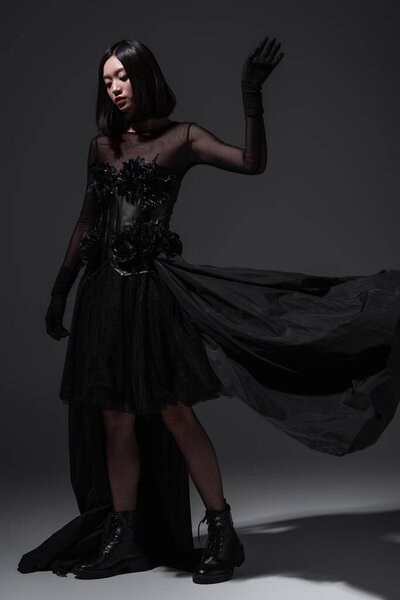 full length of young asian woman in black gothic dress and gloves posing on dark grey