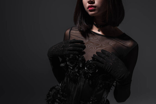 обрезанный вид брюнетки молодой женщины в черно-готическом платье и перчатки, позирующие на темно-серой