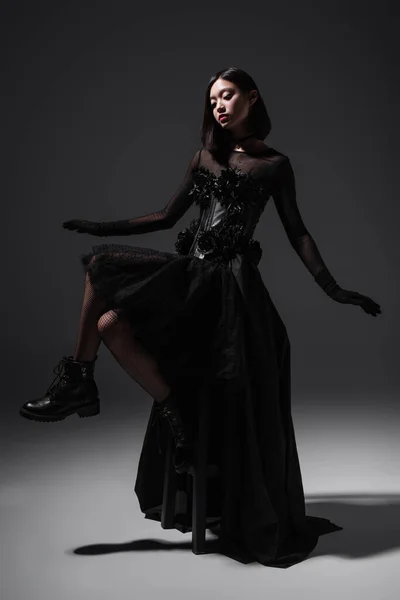 full length of asian young woman in black gothic dress and gloves sitting on high chair on dark grey