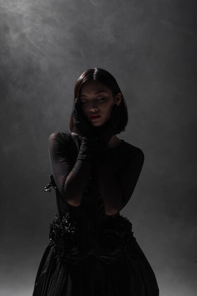 брюнетка азиатская модель с закрытыми глазами позирует в черно-готическом наряде на темно-сером с дымом