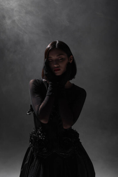 brunette asian model with closed eyes posing in black gothic outfit on dark grey with smoke