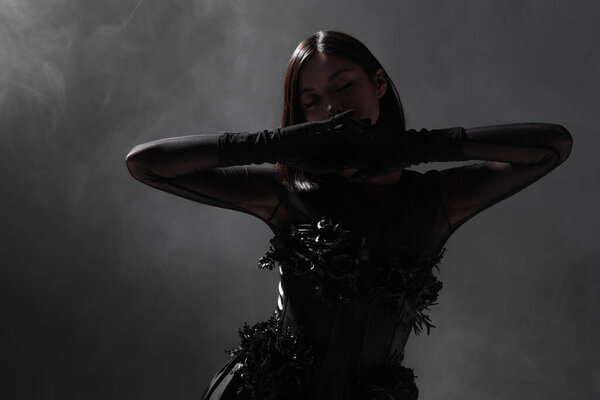 brunette asian model with closed eyes posing in gloves on dark grey with smoke