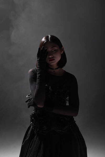 brunette asian model with closed eyes posing in black gloves and gothic outfit on dark grey with smoke