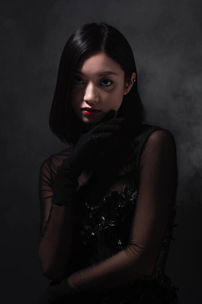 portrait of young asian woman in gothic outfit and gloves looking at camera on grey with smoke