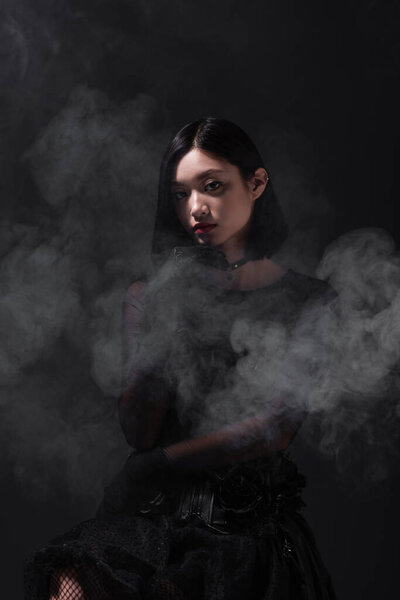 brunette young asian woman in gothic outfit and gloves on grey with smoke