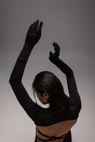 back view of brunette asian woman in total black outfit with gloves gesturing isolated on grey 