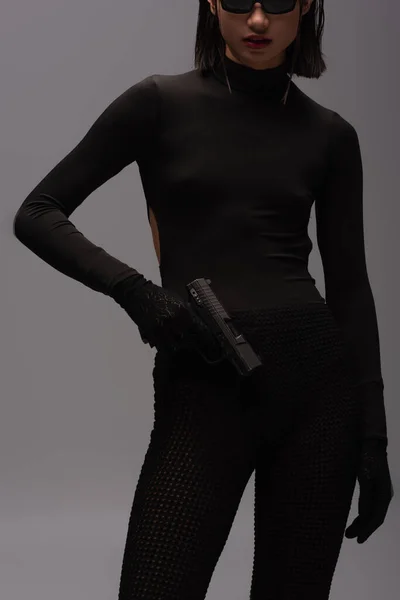 cropped view of dangerous asian woman in total black outfit and stylish sunglasses holding gun isolated on grey