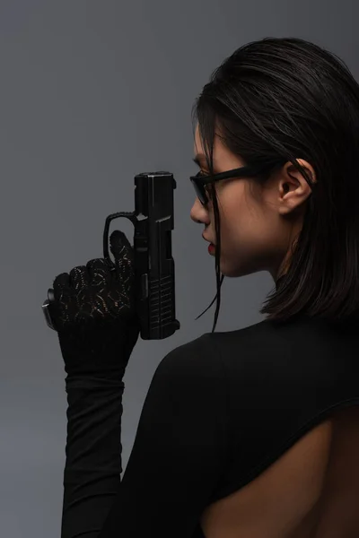 side view of dangerous asian woman in total black outfit and stylish sunglasses holding gun isolated on grey