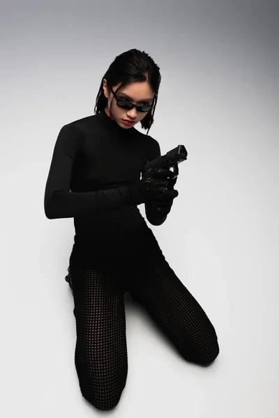 dangerous asian woman in total black outfit and stylish sunglasses holding gun on white