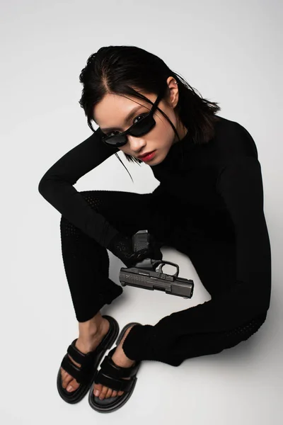 top view of asian woman in total black outfit and stylish sunglasses sitting near gun on white
