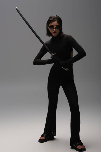 полная длина молодой азиатской женщины в солнцезащитных очках и черном наряде держа катану на сером