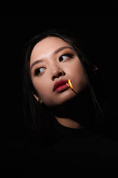 portrait of pretty asian woman with burning match in mouth looking away isolated on black