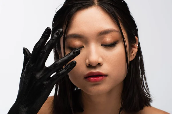 young asian woman with closed eyes and hand in black paint posing isolated on white