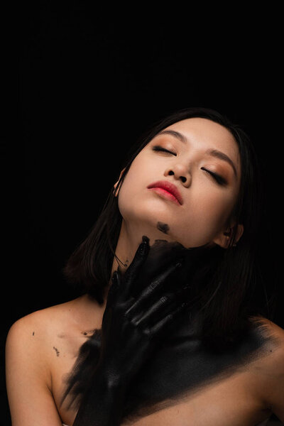 young asian woman with bare shoulders and hand in paint touching neck isolated on black
