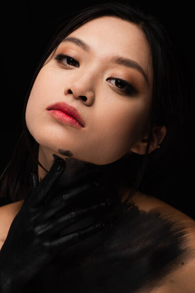 portrait of young asian woman with hand in paint touching neck isolated on black