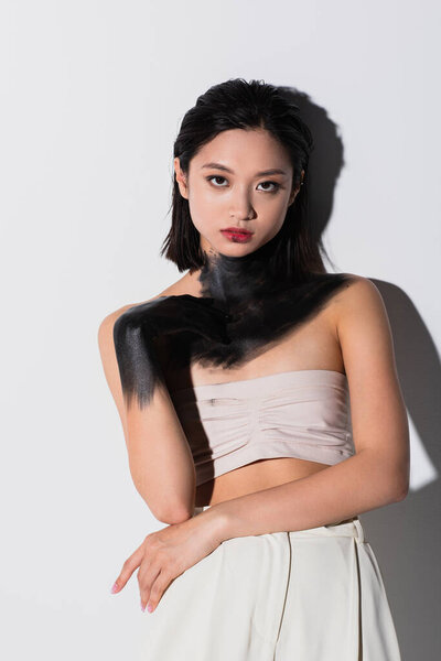 young asian woman with bare shoulders and hand in black paint looking at camera on white
